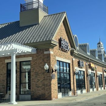 Faces by Freda - The image shows the exterior of a brick commercial building with multiple large windows and signage for Salon Freda, a beauty studio. The building features a metal roof with a gabled design and decorative architectural elements, including a small tower with a spire. There are outdoor seating arrangements near the entrance, and the area appears to be part of a shopping plaza or retail complex. The sky is clear and blue, indicating a sunny day. - Waxing Near Me - Novi, Michigan
