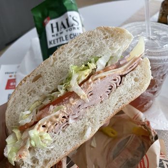 Brooklyn Bread Cafe - The Mila - A close-up of a sandwich featuring sliced turkey, shredded lettuce, tomato slices, and mayonnaise in a crusty baguette. The sandwich is placed on a paper wrapper, with a drink in a plastic cup with a straw and a bag of Halls kettle chips in the background. The setting appears to be a casual dining area or café. - Sandwiches Near Me - Brooklyn, New York