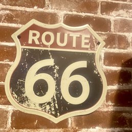 A Route 66 sign with a shield shape is mounted on a brick wall. The sign has a black background with white lettering and a red top section that reads "ROUTE." The sign appears aged, with visible scratches, paint chipping, and signs of weathering, giving it a vintage look. The brick wall behind it has a reddish-brown color with white mortar lines, adding to the rustic aesthetic.
