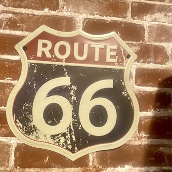 Pizza 9 - A Route 66 sign with a shield shape is mounted on a brick wall. The sign has a black background with white lettering and a red top section that reads "ROUTE." The sign appears aged, with visible scratches, paint chipping, and signs of weathering, giving it a vintage look. The brick wall behind it has a reddish-brown color with white mortar lines, adding to the rustic aesthetic. - Pizza Near Me - Grants, New Mexico