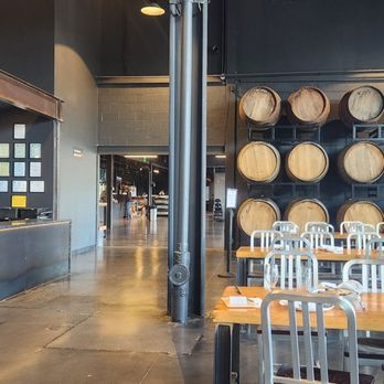 Trillium - Canton - Indoor tables for drinking and dining - The image shows an indoor dining space with a modern industrial design, including a wall decorated with multiple wooden barrels arranged on shelves. There are several wooden tables with metal chairs set up for seating. To the left, a counter with a wood storage area filled with chopped firewood is visible. The space has high ceilings with exposed beams and lighting fixtures, and a doorway leading to another area in the background. - Breweries Near Me - Canton, Massachusetts