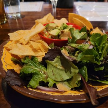 Cafecito - A ceramic plate contains a mixed green salad with various leafy greens, a small red bowl of guacamole with avocado pieces, and tortilla chips. The plate is placed on a dark wooden table, with glasses and menus visible in the background, indicating a restaurant setting. - Cuban Near Me - Chicago, Illinois