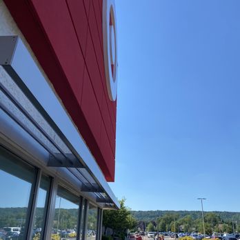 Target - The image shows the exterior of a Target retail store with a red facade and the Target logo mounted on the building. The store has large glass windows and a sidewalk in front, with parking spaces and cars visible in the background. The scene is set on a clear day with a blue sky and distant green hills. - Department Stores Near Me - Vauxhall, New Jersey