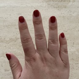 A photograph features a hand with four fingers visible, each with red nail polish. The background is a beige, textured surface, possibly a stone or tile. The hand appears to be held up in front of the camera, with the focus on the nails and fingers.