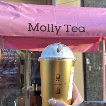 Molly Tea - Snowy White Jasmine Tea; IG: susanlamx - A person holds a plastic cup of white jasmine tea with ice, featuring a gold and white design with Japanese characters and the word 'Jianshan.' The background shows the storefront of Molly Tea with a pink awning and some outdoor elements, including a water bottle and a coffee cup on the counter. The scene is outdoors during daylight. - Bubble Tea Near Me - Brooklyn, New York