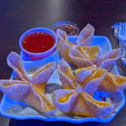 a plate of food on a table - The image shows a white, decorative plate containing several fried wontons dusted with powdered sugar. A small cup of red dipping sauce and a small container of soy sauce are placed on the plate. The plate is set on a dark table, with a blurred background. The presentation suggests a serving of Asian-style appetizers, likely at a casual dining restaurant.