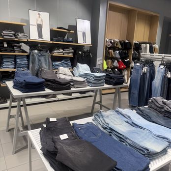 H&M-Paramus - Folded jeans and pants are arranged on tables and shelves in a retail clothing store, with racks of additional denim in the background. - Women's Clothing Near Me - Paramus, New Jersey