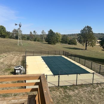 Wideman Pools - A rectangular outdoor swimming pool is covered with a dark green pool cover and surrounded by a black metal fence. The pool area is on a concrete or paved surface, with a grassy field and trees in the background. A windmill is visible on a hill in the distance, indicating a rural setting. The sky is clear and blue, suggesting a sunny day. - Hot Tub & Pool Near Me - Festus, Missouri