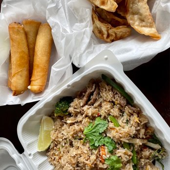 Thai Time - Basil fried rice - The image shows a Thai meal with a container of fried rice garnished with cilantro, lime, and vegetables, along with two spring rolls and fried dumplings served in separate paper-lined baskets. The fried rice includes broccoli, carrots, and green onions, and is presented in a white takeout container. The spring rolls are golden brown, and the fried dumplings are crispy with a slightly browned exterior. The food is arranged on a dark table surface. - Food Trucks Near Me - Austin, Texas