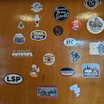 Three Falls Cider - Door with stickers - The image shows a wooden wall adorned with numerous stickers representing breweries, cideries, outdoor parks, and related themes. Notable stickers include logos for Woodlawn Distilling, Aurora Brew Works, Silver Lake Brewing Project, and North High Brewing. There are also stickers for local parks like Letchworth State Park, and various decorative or humorous designs, such as a bee with the message 'Save the Bees' and a cartoon character. The stickers are arranged in a scattered pattern across the wooden surface. - Cideries Near Me - Castile, New York