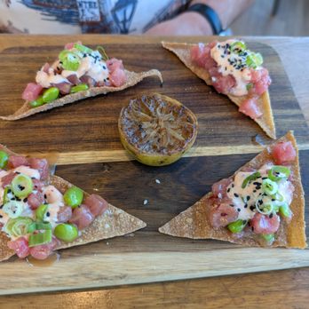 Devil's Kitchen - Tuna Tostadas - Four tuna tostadas are arranged on a rectangular wooden serving board, each topped with diced tuna, sliced green onions, edamame, black sesame seeds, and a creamy sauce. A grilled lime half is placed in the center of the board, adding a citrus element to the presentation. The background shows part of a person's arm and a wooden table surface. - New American Near Me - Grand Junction, Colorado