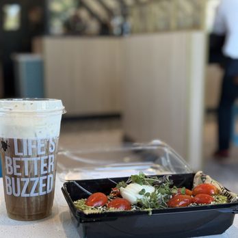 Better Buzz Coffee Tempe Market Station - This is the avocado toast with egg as well as the pumpkin cream cold brew I love the backdrop. - A black takeout container holds a salad with cherry tomatoes, microgreens, and cheese, placed on a table at a cafe. A cold brew coffee with foam and a straw is nearby. - Cafes Near Me - Tempe, Arizona