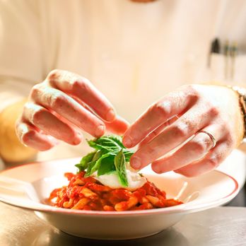 Felice 56 - A close-up action shot of a chef finishing a dish of pasta topped with tomato sauce and a creamy ball of burrata, adding a fi... - Italian Near Me - New York, New York Felice 56 - A close-up action shot of a chef finishing a dish of pasta topped with tomato sauce and a creamy ball of burrata, adding a fi... - Italian Near Me - New York, New York
