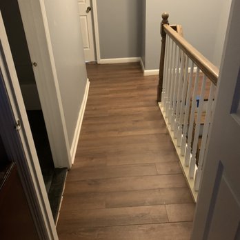 Munoz Construction - Hallway in progress - The image shows a hallway with wood laminate flooring leading to a staircase on the right side, which has white balusters and a wooden handrail. The walls are painted light gray, with a framed picture hanging on the wall near the door. There is a closed door at the end of the hallway, and a light switch is visible on the wall. The space appears clean and well-maintained, with baseboards along the walls. - General Contractors Near Me - Herndon, Virginia