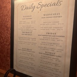 A printed menu board titled 'Daily Specials' lists meal options for Tuesday through Sunday, including soups, sandwiches, and dinners. Each day features specific dishes such as beef stew, chicken vegetable soup, and seafood platters, with prices and descriptions. The board is framed and placed on a wooden surface, with a warm, inviting background.