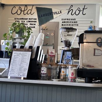 Meta Cafe - The photo depicts the interior counter of Meta Cafe, including coffee brewing equipment, a menu stand, a decorative potted plant, and containers of tea. A wall sign behind the counter displays beverage options like espresso, Americano, and macchiato, with the words 'cold' and 'hot' written above. The scene includes a window on the right, allowing natural light into the space, and various coffee-related items arranged on the counter. - Sandwiches Near Me - Lambertville, New Jersey
