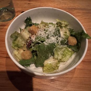 Prato - Escarole Caesar - A ceramic bowl contains a Caesar salad with mixed leafy greens, croutons, and shredded cheese on top. The bowl is on a wooden table, with a glass of water and a plate with a spoon visible in the background. The setting appears to be a restaurant or dining area. - Pizza Near Me - Winter Park, Florida