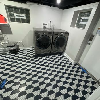 MDF Construction - A laundry room features a pair of front-loading washer and dryer, wall-mounted shelves with cleaning supplies, and a black-and-white geometric patterned floor. - Painters Near Me