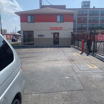 Public Storage - Closed sundays - The image shows a small, two-story building with a sign reading 'RENTAL OFFICE' on the front. The building has a beige lower section and a red upper section, with two windows on the second floor. The parking lot in front has marked parking spaces, some of which are empty. A vehicle is partially visible on the left side of the image, and a gated area with orange posts is on the right. The background includes a larger multi-story building and a clear sky with some clouds. - Self Storage Near Me - Chicago, Illinois