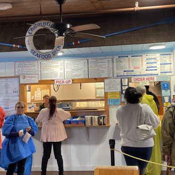 Champlin's Seafood - Order window - Several customers are standing in line at a seafood restaurant counter, with some engaging with staff behind the counter. The menu is displayed above, and signs indicate pick-up and order areas. The setting appears to be indoors with a ceiling fan and a life preserver decoration. People are dressed in casual clothing, and some are wearing jackets or raincoats. - Seafood Near Me - Narragansett, Rhode Island