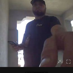 A person is standing at the front door, taking a selfie with a smartphone. The individual is smiling and wearing a cap and dark clothing. The background shows a bright outdoor area with some architectural features. The image appears to be a screenshot from a security or doorbell camera, with a timestamp and notification indicating a missed ring.