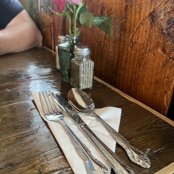 Nonna Lisas Kitchen - Fresh roses and utensils - A fork, knife, and spoon with ornate handles are placed on a folded white napkin on a dark, polished wooden table. Behind the utensils, there is a small glass jar holding a pink rose with green leaves, and a salt shaker with a metal lid. The setting appears to be in a restaurant or cafe, with a wooden panel wall and a window in the background. - Italian Near Me - Plattsburgh, New York