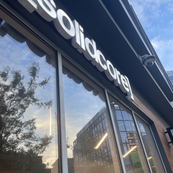 Solidcore - outside - The photo captures the exterior of 'Lolidicure,' a business with large glass windows that reflect the sky, trees, and surrounding buildings. The sign with the business name is mounted above the windows, and the image is taken from a low angle, showing part of the building's facade and the blue sky with some clouds. The reflection in the windows includes a multi-story building and trees, indicating an urban setting. - Pilates Near Me - Boston, Massachusetts