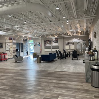 MSI - the inside of a furniture store - The image shows a spacious, modern interior with a high ceiling featuring exposed ductwork and industrial lighting. The floor transitions from wood to tile, with seating arrangements including chairs, a sofa, and tables. Display samples or materials are mounted on the walls, and there are decorative elements like string lights and framed pictures. The space appears to be a showroom or retail store, with a front entrance visible in the background and various display items along the walls. - Building Supplies Near Me - Hatboro, Pennsylvania