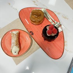 Biscoff Cheesecake - Three desserts are displayed on pink plates: a caramel pastry, a chocolate cake with raspberries, and a cannoli, set on a white table with a beige napkin.
