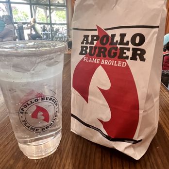 Apollo Burger - A paper bag and a plastic cup with the Apollo Burger logo are placed on a wooden table inside a restaurant. - Burgers Near Me - Lehi, Utah