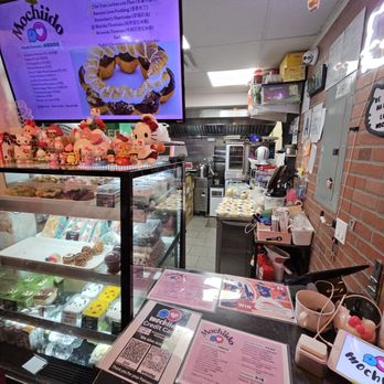 Mochiido - The image depicts the interior of Mochiido, a dessert shop specializing in Asian fusion bakery treats. A glass display case on the left contains various desserts, including macarons and other confections. The counter has menus and promotional materials, including a pink menu and a QR code for credit card payments. The background shows the kitchen area with equipment and supplies. Decorations, such as hanging plush toys and small figurines, are visible above the display case. The space has a cozy, cluttered appearance with a brick wall on the right and a ceiling with suspended decorations. - Donuts Near Me - Flushing, New York