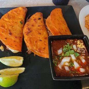 Mezcalitos - Birria tacos - Three birria tacos served on a black plate with lime wedges and a small bowl of consommé, accompanied by rice on a white plate. - Mexican Near Me - Cedar Falls, Iowa