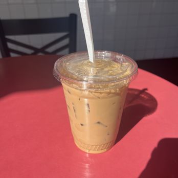 Saigon Express - Saigon Coffee - A transparent plastic cup contains iced coffee with visible ice cubes and a light brown color, topped with a clear plastic lid and a white straw. The cup is on a red table, with sunlight casting a shadow of the cup and straw. The background includes a chair and a tiled wall, suggesting an outdoor or cafe setting. - Vietnamese Near Me - Berkeley, California
