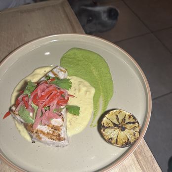 Osa Rooftop - Grilled Swordfish was surprisingly incredible with corn and ajo verde sauce - A dish featuring grilled swordfish topped with pickled vegetables, accompanied by green sauce and grilled lemon, on a beige plate. A dog is visible in the background. - Bars Near Me - Pasadena, California