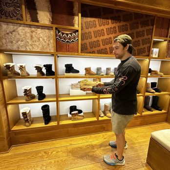 UGG - A man with a cap, black long-sleeve shirt, and shorts is standing in front of a wooden display shelf filled with various UGG boots. He is holding a pair of boots and appears to be examining them. The store has wooden flooring and a decorative wooden wall with UGG branding in the background. - Men's Clothing Near Me - East Rutherford, New Jersey