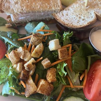 Milkstand - Turkey Club Sandwich - A plate features a turkey club sandwich made with toasted bread, layered with turkey, lettuce, and possibly other fillings. Accompanying the sandwich is a side salad consisting of mixed greens, shredded carrots, croutons, cucumber slices, and tomato wedges. A small container of dressing is also visible on the plate, suggesting a meal served at a casual dining establishment. - Breakfast & Brunch Near Me - Ithaca, New York