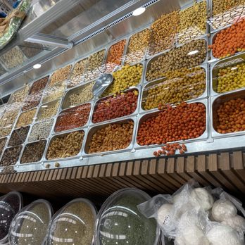 Jerusalem Roastery - Mix nuts roasted in house. - Multiple metal containers are arranged on a store shelf, each filled with different dried fruits, nuts, and seeds in various colors including orange, yellow, brown, and beige. A metal scoop is placed in one of the containers, indicating self-service. Below the shelf, clear plastic containers hold additional dried goods. The setting appears to be a specialty store offering a variety of dried foods and ingredients. - Candy Stores Near Me - Garden Grove, California