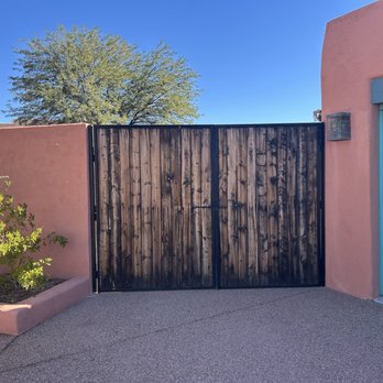 First Impression Ironworks - Old gate - The image features a dark wooden gate with vertical planks, framed by pink stucco walls on either side. To the right, part of a blue garage door is visible, and a small outdoor light fixture is mounted on the wall near the gate. A large tree with green foliage is seen in the background against a clear blue sky, and a small cactus plant is situated in a pink planter bed on the left side of the image. The ground is paved with concrete. - Fences & Gates Near Me - Gilbert, Arizona