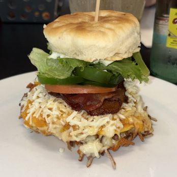 11 Degrees North - Cheesie - A burger features shredded cheese, bacon, fresh lettuce, tomato slices, and jalapeño peppers, all assembled on a plate. The burger is topped with a soft bun secured with a toothpick, and the background shows a drink and condiments, indicating a casual dining setting. - Coffee & Tea Near Me - Chicago, Illinois