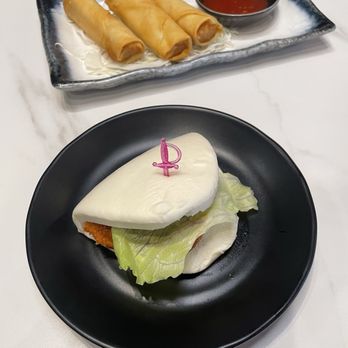 Ichiddo Ramen - A bao bun with a pink candle on top is placed on a black plate, containing a leaf of lettuce and a fried filling. Behind it, a rectangular ceramic dish holds four spring rolls with a small bowl of red dipping sauce. The setting appears to be a restaurant table with a white surface. - Ramen Near Me - Fort Wayne, Indiana