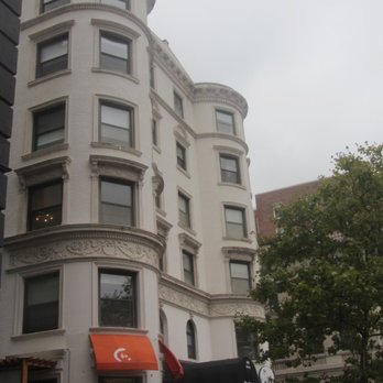 Kenmore Square - A multi-story building featuring ornate architectural details, including decorative moldings and rounded corner towers, located on a city street. The ground level has storefronts with signage, including an orange awning and an open sign, and a tree partially obscures part of the building. The sky is overcast. - Landmarks & Historical Buildings Near Me - Boston, Massachusetts