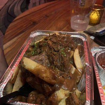 Devil's Hideaway - Poutine fries - A foil tray contains poutine fries topped with beef gravy and garnished with chopped green herbs. The tray is placed on a wooden table, with a plastic cup of water, a small container of sauce, and other dining items visible in the background. The setting appears to be a casual dining environment, possibly a restaurant or bar. - Wine Bars Near Me - Tempe, Arizona
