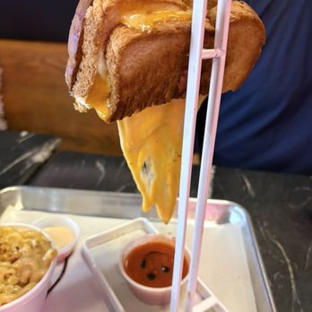 Grilled Cheese Gallery - Best grilled cheese ever - A grilled cheese sandwich with melted cheese is displayed on a metal stand, with dipping sauces and macaroni and cheese on a tray. - Ice Cream & Frozen Yogurt Near Me - St. Augustine, Florida