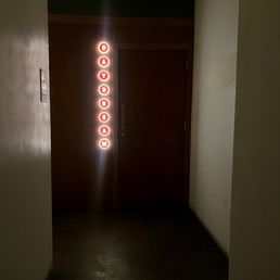 Secret entrance - A dark corridor leads to a closed wooden door, with a vertical sign to the left of the door displaying the word 'DAYDRIAM' in red, illuminated letters. The sign is composed of individual circular lights with each letter inside. The surrounding area is dimly lit, with plain walls and a concrete floor, suggesting an indoor setting.
