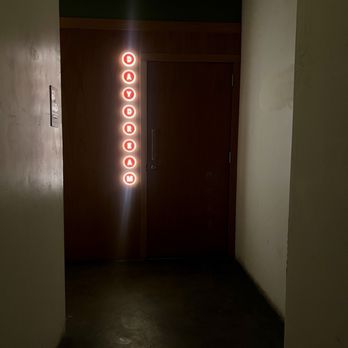 Daydream - Secret entrance - A dark corridor leads to a closed wooden door, with a vertical sign to the left of the door displaying the word 'DAYDRIAM' in red, illuminated letters. The sign is composed of individual circular lights with each letter inside. The surrounding area is dimly lit, with plain walls and a concrete floor, suggesting an indoor setting. - Cocktail Bars Near Me - Albuquerque, New Mexico