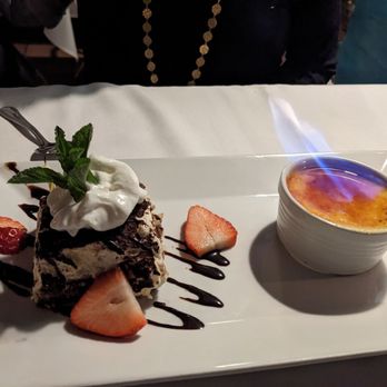 Ristorante Euno - Birthday desserts: Flaming Crème Brulee and Tiramisu - The image shows a rectangular white plate with two desserts: a chocolate cake topped with whipped cream, a mint leaf, and strawberries, decorated with chocolate sauce; and a ramekin of crème brûlée with a caramelized sugar crust. The plate is on a white tablecloth, and part of a person wearing dark clothing and a gold necklace is visible in the background. - Italian Near Me - Boston, Massachusetts