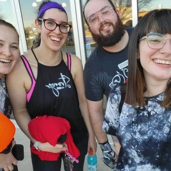 KickHouse - A group of four people, three women and one man, are standing close together outdoors in front of a glass door, smiling at the camera. They are dressed casually, with some wearing glasses and one holding a water bottle. The background shows reflections of trees and the sky in the glass, indicating a sunny day. - Gyms Near Me - Chantilly, Virginia