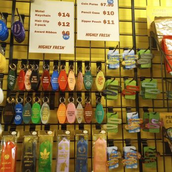 Highly Fresh - Keychains, ribbons and hair clips. - The image shows a retail display on a yellow grid wall holding various novelty keychains, ribbons, and hair clips. The keychains have humorous and colorful designs, each attached to hooks with price tags. Two white signs list prices for items such as motel keychains, hair clips, award ribbons, and accessories like coin purses and pencil cases. The display is organized with different types of keychains and accessories, some with themed or humorous messages, and others with decorative elements. The overall setting appears to be a gift or souvenir shop. - Gift Shops Near Me - Ottawa, Ontario