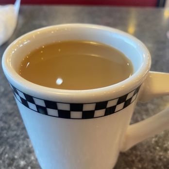 Big Easy Diner - Nice cup of coffee - A white ceramic mug with a black checkered pattern near the rim contains coffee and is positioned on a dark, speckled countertop. The mug has a handle on the right side, and the coffee appears to be light brown. The background shows a blurred red booth or seating area, indicating a casual dining or cafe setting. - American Near Me - Slidell, Louisiana