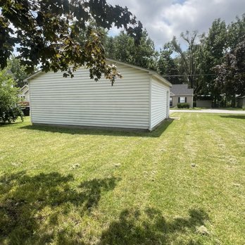 Hope Cleaning - The image shows a white shed with vinyl siding located on a grassy lawn. The shed is rectangular with a sloped roof and is positioned near the center of the yard. Surrounding the shed are trees providing partial shade, with some shadows cast on the grass. In the background, residential houses and a street are visible, along with a partly cloudy sky overhead. - Home Cleaning Near Me